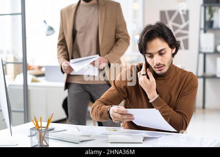 Giovane uomo d'affari che legge contratto mentre ha una conversazione sul telefono mobile, lui lavora al suo posto di lavoro in ufficio Foto Stock