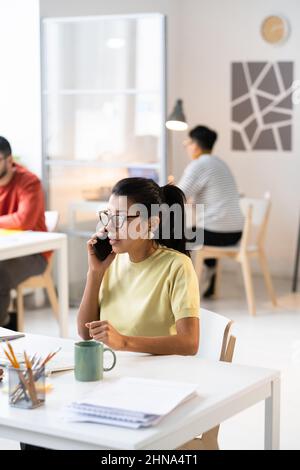 Donna d'affari in occhiali concentrandosi sulla chiamata seria sul telefono mobile mentre si siede al suo posto di lavoro in ufficio Foto Stock