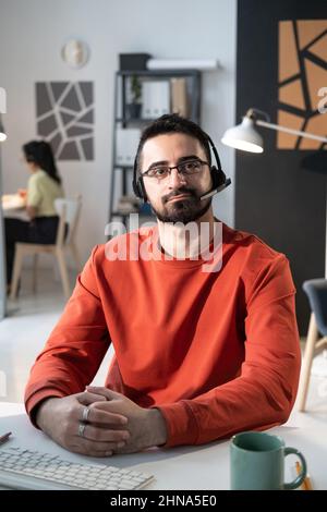 Ritratto dell'operatore del servizio clienti in occhiali e cuffie che guarda la fotocamera mentre lavora con i clienti in ufficio Foto Stock