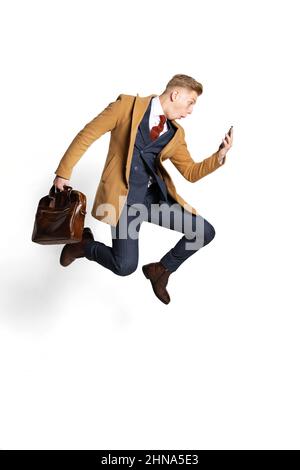 Sorpreso giovane uomo d'affari, studente, diplomatico saltando con smartphone isolato su sfondo bianco studio. Emozioni umane, concetto di espressione facciale. Foto Stock