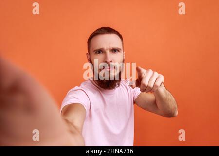 Ritratto di un uomo orrido serio e bello che fa selfie o streaming, puntando alla fotocamera, POV, punto di vista della foto, indossando una T-shirt rosa. Studio interno girato isolato su sfondo arancione. Foto Stock