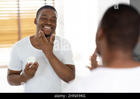 Cura post-rasatura. Attraente ragazzo Afro-Americano usando crema idratante in bagno Foto Stock