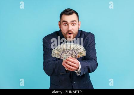 Avido uomo d'affari che indossa tuta di stile ufficiale che tiene molto denaro ed esprime l'eccitazione, mostrando bollette del dollaro, sorpreso di successo finanziario. Studio interno girato isolato su sfondo blu. Foto Stock