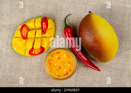 Vista dall'alto di mango intero, peperoncino intero, pepe caldo tagliato a fette, metà di mango tagliata e chutney di mango piccante in un recipiente di vetro sul burlap. Foto Stock