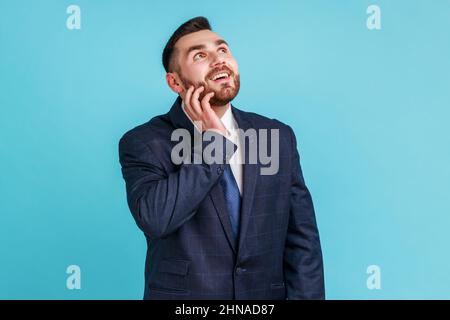 Sorridente uomo adulto che indossa un abito ufficiale, con volto tranquillo pensando ai piani per il futuro tenere il mento, sognando di buon lavoro. Studio interno girato isolato su sfondo blu. Foto Stock