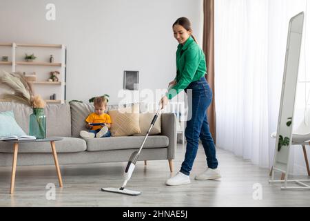 Felice madre giovane mordere il pavimento e riordinare mentre suo figlio seduto sul divano e guardare cartoni animati sullo smartphone Foto Stock