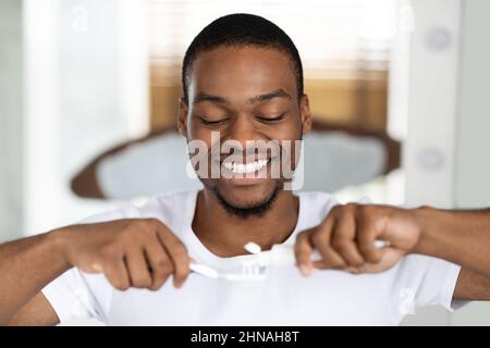 Ritratto di Happy Black Guy spremitura dentifricio su spazzolino in bagno Foto Stock