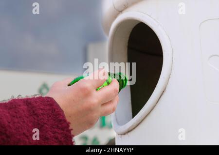 Una donna o una donna che gettano una bottiglia di vetro verde vuota nel contenitore per rifiuti del cestino. Amore natura, cernita rifiuti, riciclaggio, concetto di cura idea. Foto Stock