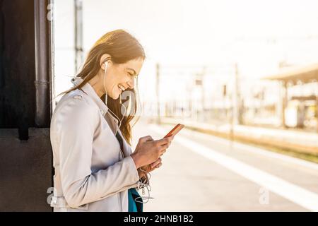 Donna d'affari sorridente usando lo smartphone mentre aspettava in una stazione ferroviaria - ragazza giovane e attraente studentessa usando il cellulare mentre in piedi Foto Stock