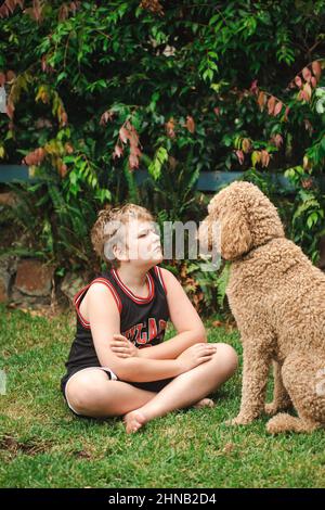 Ragazzo pre-teen seduto su erba giocando con grande cane buon Doodle d'oro Foto Stock