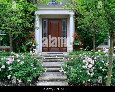 Casa e giardino con cespugli di rose e scalini in pietra che conducono all'elegante porta anteriore in grano di legno Foto Stock