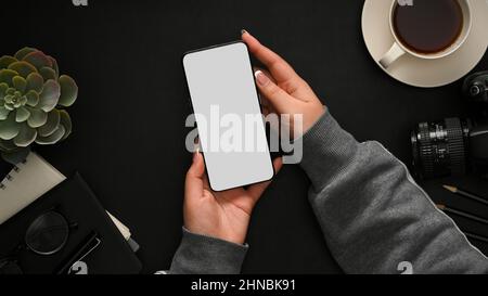 Donna che utilizza il telefono cellulare al tavolo nero dell'ufficio con forniture per ufficio e tazza di caffè. Vista dall'alto spazio di lavoro nero e smartphone con schermo vuoto. Foto Stock