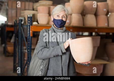 Donna anziana in maschera protettiva alla ricerca di vasi di fiori in negozio Foto Stock