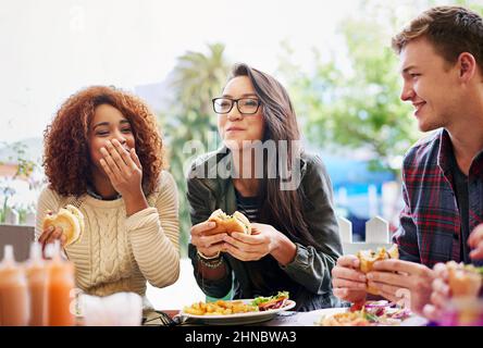 Buon cibo e risate vanno di pari passo. Scatto corto di tre amici che mangiano hamburger all'aperto. Foto Stock