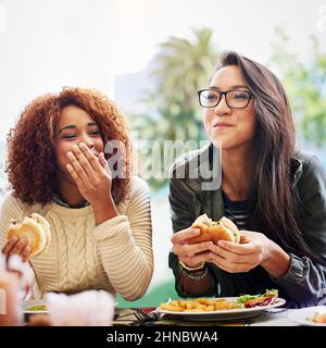 Condividere il buon cibo e fare delle battute. Scatto corto di due girlfriends che mangiano gli hamburger all'aperto. Foto Stock