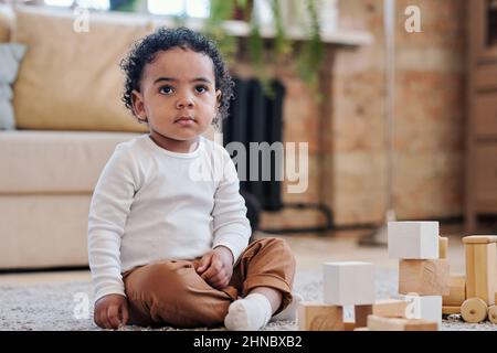 Bimbo nero concentrato con capelli ricci seduti su tappeto e giocando con i soli giocattoli Foto Stock