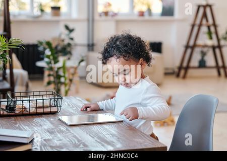 Toddler da corsa misto moderno concentrato con capelli ricci in piedi alla scrivania e utilizzando il tablet in salotto Foto Stock
