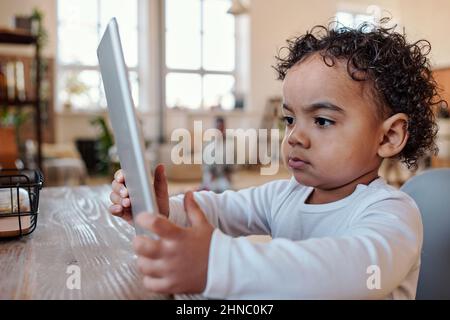 Bimbo afro-americano concentrato con capelli ricci seduti al tavolo e con l'app tablet Foto Stock