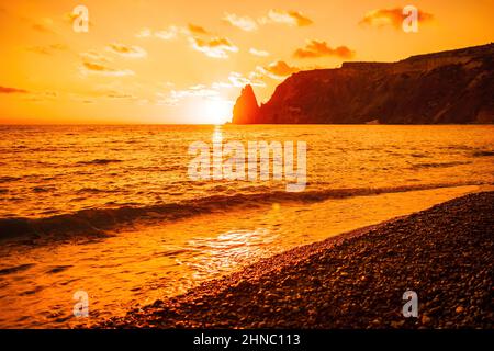 Un tramonto rosso acceso sul mare con scogliera vulcanica rocciosa. Astratta natura estate o primavera oceano mare sfondo. Piccole onde su acqua calda dorata Foto Stock