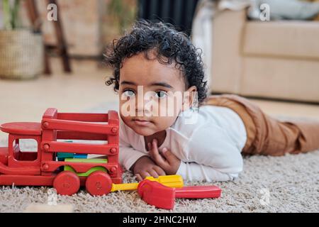 Ritratto di ragazzo nero con capelli ricci seri che giace vicino a un'auto giocattolo su tappeto in soggiorno Foto Stock