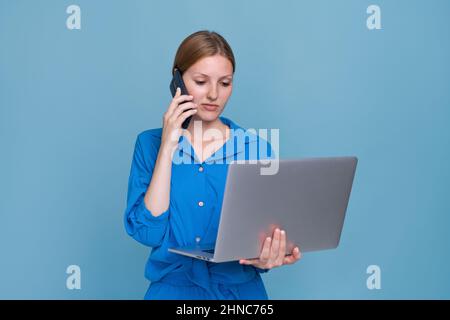 Ritratto moderno felice sorridente giovane donna che chiama su smartphone con notebook su sfondo blu in una camicia blu. Ragazza caucasica libera chiama a lavorare con un cliente negoziazione Foto Stock