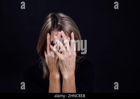 Giovane donna infelice in blusa nera con mani che coprono il viso, occhi che guardano la fotocamera attraverso le dita su sfondo nero. Fisico e psicologico Foto Stock