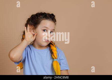 Curioso interessato piccola ragazza caucasica che hanno trecce kanekalon con occhi luminosi che tengono le orecchie per ascoltare qualcosa che guarda la macchina fotografica sul beige Foto Stock