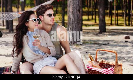 Giovane donna in occhiali da sole che tiene il vino vicino al ragazzo e cestino sulla spiaggia Foto Stock