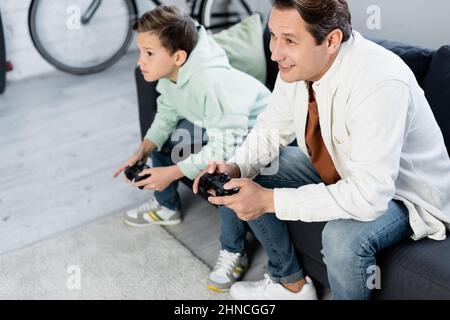 KIEV, UCRAINA - 11 OTTOBRE 2021: Uomo sorridente che gioca video game con figlio sfocato a casa Foto Stock