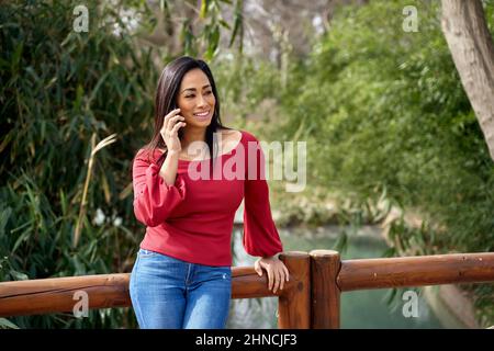 Sorridente femmina ispanica che parla al telefono mentre si trova in piedi sul ponte sopra lo stagno vicino agli alberi verdi nel parco il giorno d'estate Foto Stock