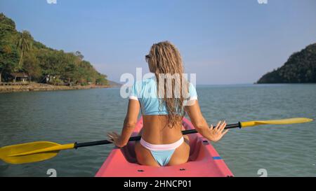 Lunga donna bionda ricci capelli in fila di costume da bagno kayak di plastica lungo azzurro oceano acqua presso il resort esotico vicino retro vista. Viaggiare in paesi tropicali. La ragazza sta navigando in kayak nell'oceano. Foto Stock