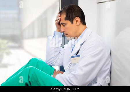 Scatto corto di un bel giovane medico maschio con un mal di testa che tiene la testa mentre si appoggia contro un muro nel suo ospedale Foto Stock