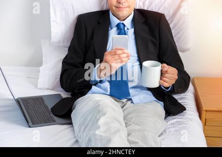 Uomo d'affari maturo che tiene una tazza di caffè e lavora con lo smartphone a letto Foto Stock