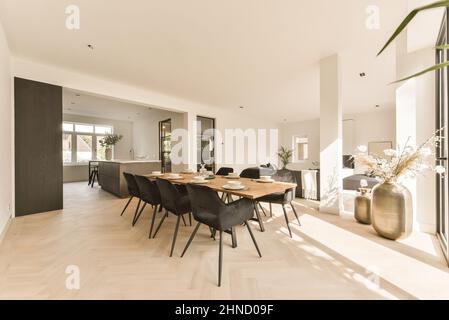 Sedie a tavola in legno con stoviglie poste in sala da pranzo vicino zona cucina in spazioso appartamento con fiori in vaso Foto Stock