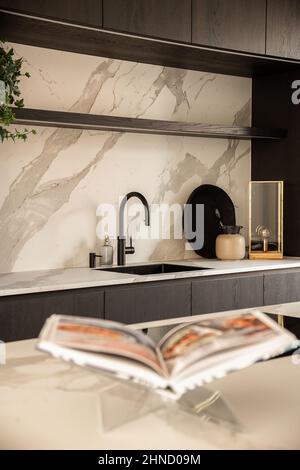 Bancone con libro aperto su stand contro lavandino con dispenser e armadi neri in luminosa cucina moderna di elegante appartamento Foto Stock