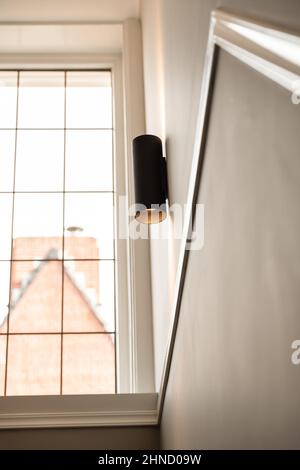 Da sotto di elegante lampada nera appesa su una parete grigia vicino a una grande finestra di vetro nel corridoio luminoso dell'appartamento moderno Foto Stock