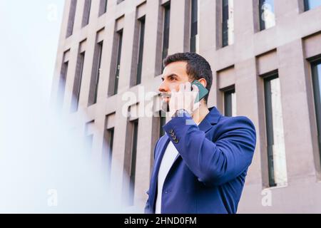 Da sotto contenuto portava imprenditore maschile in abbigliamento formale guardando via mentre si ha conversazione telefonica vicino moderno edificio a più piani su strada di Foto Stock
