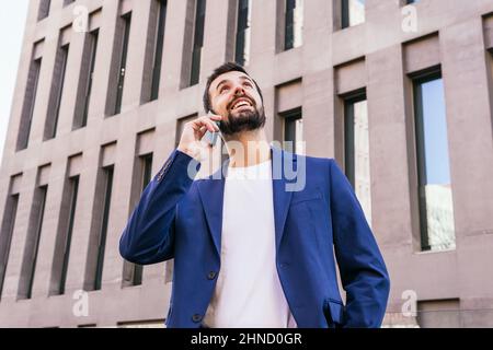 Dal contenuto di sotto ha bearded imprenditore maschile in abbigliamento formale guardando in su, mentre avendo conversazione telefonica vicino moderno edificio a più piani sulla strada di ci Foto Stock