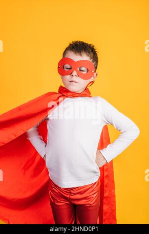 Capretto coraggioso in capo supereroe rosso e maschera in piedi sullo sfondo giallo con le mani sulla vita Foto Stock