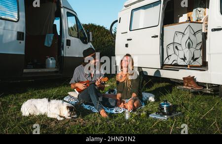 Corpo pieno di hipster positivo Donna con gli occhi chiusi ascoltando l'uomo che suona la canzone su ukulele mentre con il cane e gatto vicino ai furgoni campeggio durante la strada tri Foto Stock