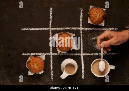 Vista dall'alto della persona anonima del raccolto che aggiunge zucchero nella tazza con caffè con latte sulla griglia del gioco Tic tac TOE con muffin dolci su sfondo nero Foto Stock