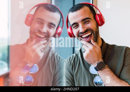 Maschio felice con la barba che ascolta la musica in cuffie wireless rosse mentre si trova vicino alla finestra con riflessi nella stanza della luce Foto Stock