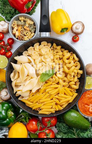 Dalla parte superiore della padella con vari tipi di pasta cruda circondata da verdure crude assortite su tavola di marmo in cucina leggera Foto Stock