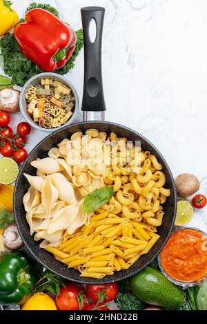 Dalla parte superiore della padella con vari tipi di pasta cruda circondata da verdure crude assortite su tavola di marmo in cucina leggera Foto Stock