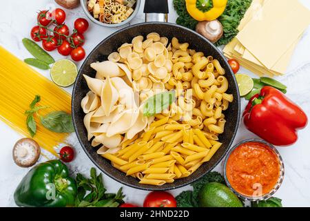 Dalla parte superiore della padella con vari tipi di pasta cruda circondata da verdure crude assortite su tavola di marmo in cucina leggera Foto Stock