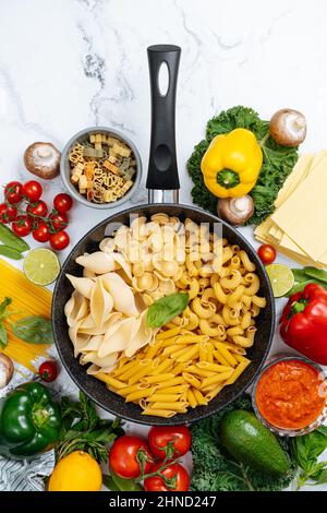 Dalla parte superiore della padella con vari tipi di pasta cruda circondata da verdure crude assortite su tavola di marmo in cucina leggera Foto Stock