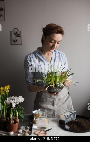 Content female gardener in apron standing with assorted potted fresh flowers and plants at table at home and looking down Foto Stock