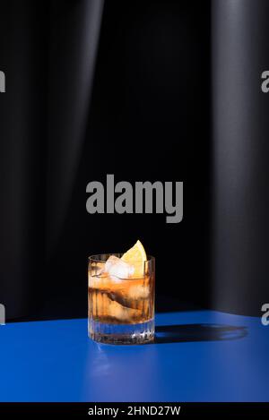 Bicchiere di cocktail alcolico preparato con vermouth rosso servito con fette d'arancia in studio chiaro con pareti blu e nere Foto Stock
