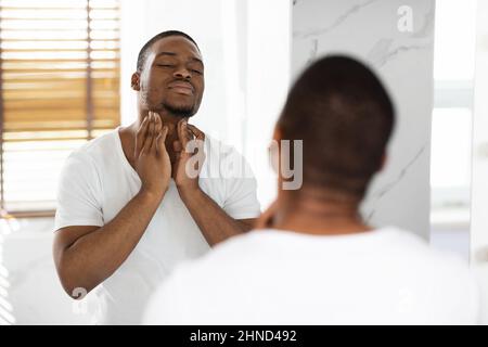 Concetto di assistenza sanitaria. Preoccupato uomo nero che tocca il collo, esaminando i linfonodi vicino a Mirror Foto Stock