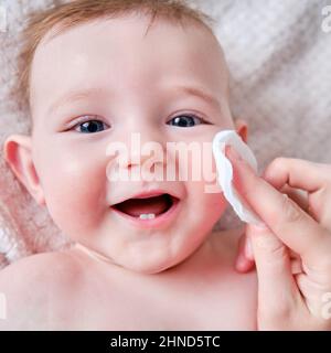 Le mani della madre lavano il viso di un bambino felice con un batuffolo di cotone. La mamma tiene il bambino sorridente dell'igiene sul divano, sei - sette mesi Foto Stock